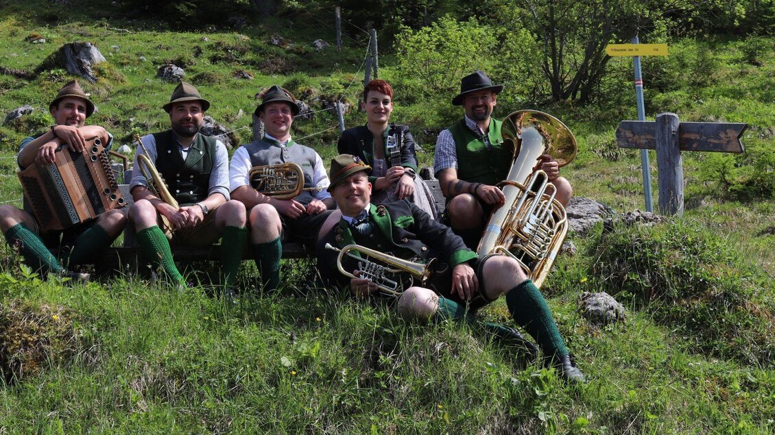 Die Musikanten von D'Röthelstoana in traditioneller Tracht  mit ihren Instrumenten der klassischen Volksmusik sitzen auf einer sommerlichen Wiese im Ausseerland.  | © Das Schutzhaus / D'Röthelstoana