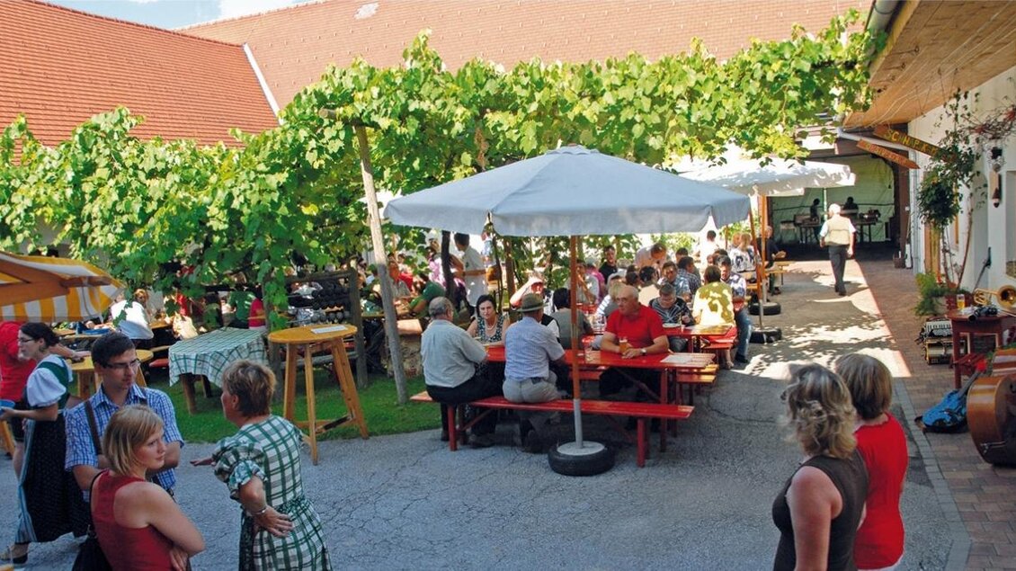 Ein belebter Garten mit Gästen, die an Tischen unter Sonnenschirmen sitzen. Umgeben von Weinreben und einem einladenden Ambiente. | © Biohof Schloffer
