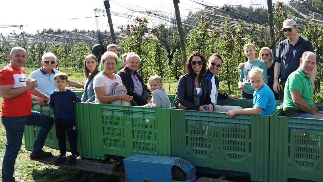 Eine Gruppe von Menschen sitzt auf einem Fahrzeug in einem Obstgarten. Im Hintergrund sind Obstbäume und eine sonnige Landschaft sichtbar. | © Biohof Schloffer