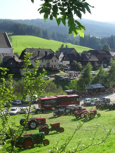 Eine malerische Landschaft mit einem kleinen Dorf und einer Kirche. Viele Traktoren und Fahrzeuge sind auf einer Wiese geparkt.
