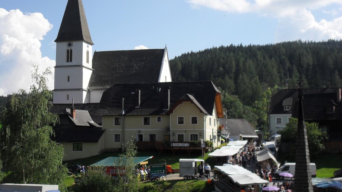 Ein malerisches Dorf mit einer Kirche im Hintergrund und einem lebhaften Markt. Die Umgebung ist von grünen Wäldern und schönen Wolken am Himmel umgeben. | © Gemeinde Hirschegg-Pack