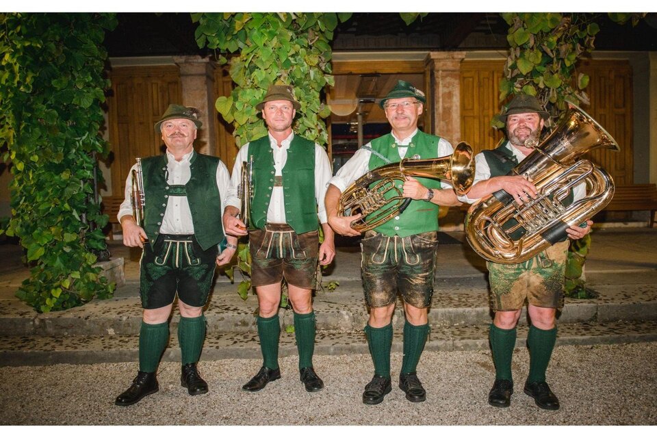 Vier Musiker in traditioneller bayerischer Tracht stehen in Reihe. Sie spielen verschiedene Blasinstrumente vor einem historischen Hintergrund. | © G.Baumann