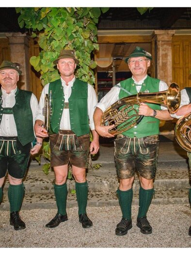 Four musicians in traditional Bavarian costume stand in a row. They play various wind instruments against a historical background. | © G.Baumann