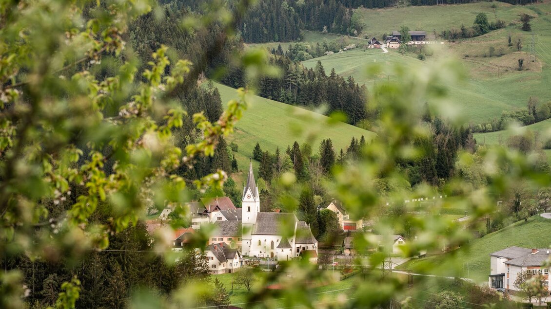 Ein malerisches Dorf umgeben von sanften Hügeln und dichten Wäldern. Im Vordergrund sind grüne Blätter sichtbar, die einen Teil der Szenerie verdecken. | © Region Graz-Mias Photoart
