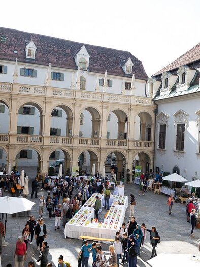 Ein belebter Innenhof mit vielen Menschen und Ständen. Sonnenschirme und historische Architektur prägen die Szene. | © c_Oliver_Wolf