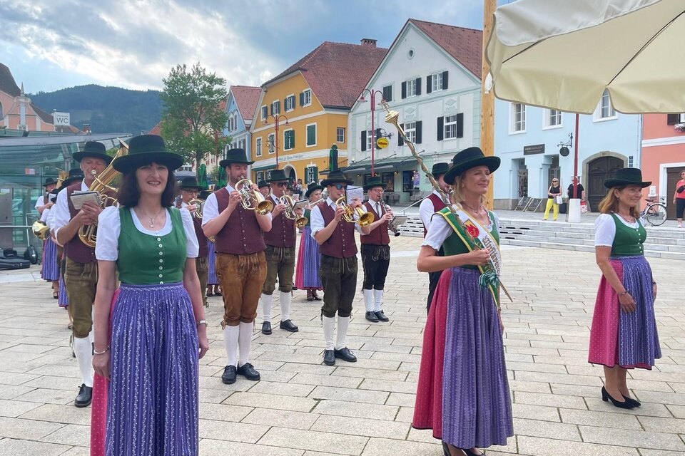 Eine Gruppe von Musikern in traditioneller Tracht spielt auf einem Platz. Im Hintergrund sind bunte Häuser und ein bewölkter Himmel zu sehen. | © (c) WOKA