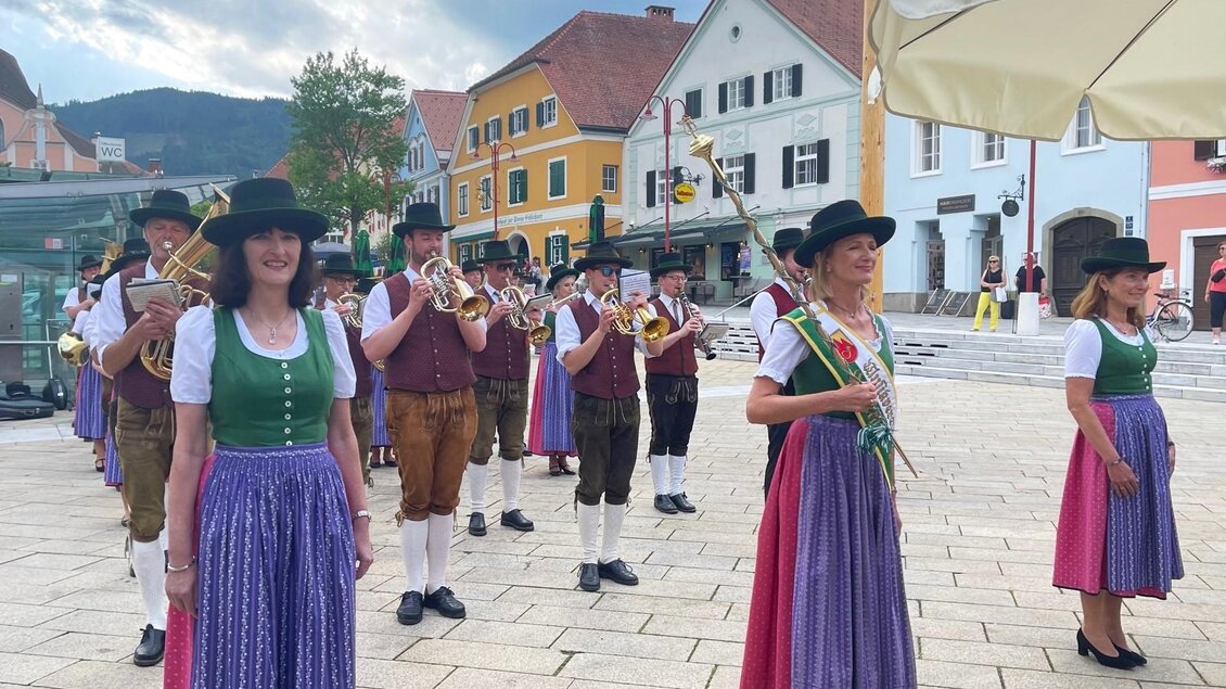 Eine Gruppe von Musikern in traditioneller Tracht spielt auf einem Platz. Im Hintergrund sind bunte Häuser und ein bewölkter Himmel zu sehen. | © (c) WOKA