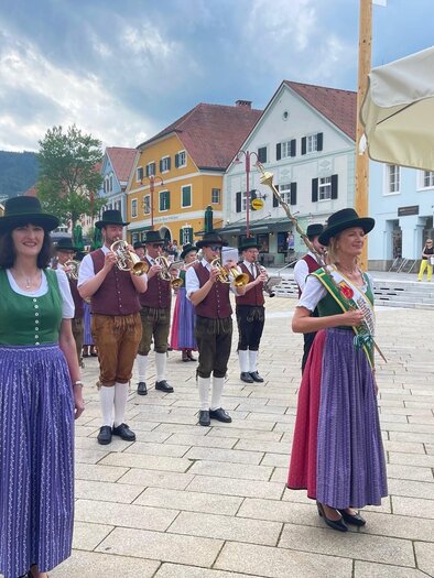 Eine Gruppe von Musikern in traditioneller Tracht spielt auf einem Platz. Im Hintergrund sind bunte Häuser und ein bewölkter Himmel zu sehen. | © (c) WOKA
