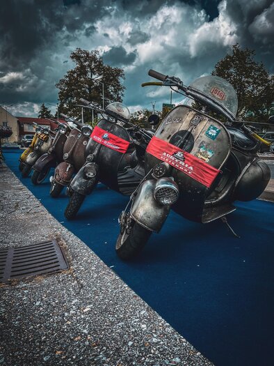 Eine Reihe alter Mopeds steht auf einer blauen Fläche. Der Himmel ist bewölkt, und die Mopeds sind mit roten Bändern dekoriert. | © Vespaclub Fürstenfeld