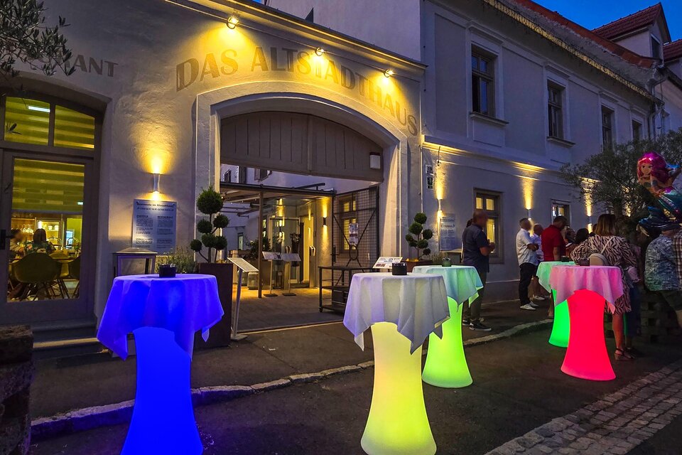 Ein Restaurant mit stilvollen, beleuchteten Stehtischen in verschiedenen Farben. Es ist Abend und Gäste stehen vor dem Eingang. | © © Das Altstadthaus