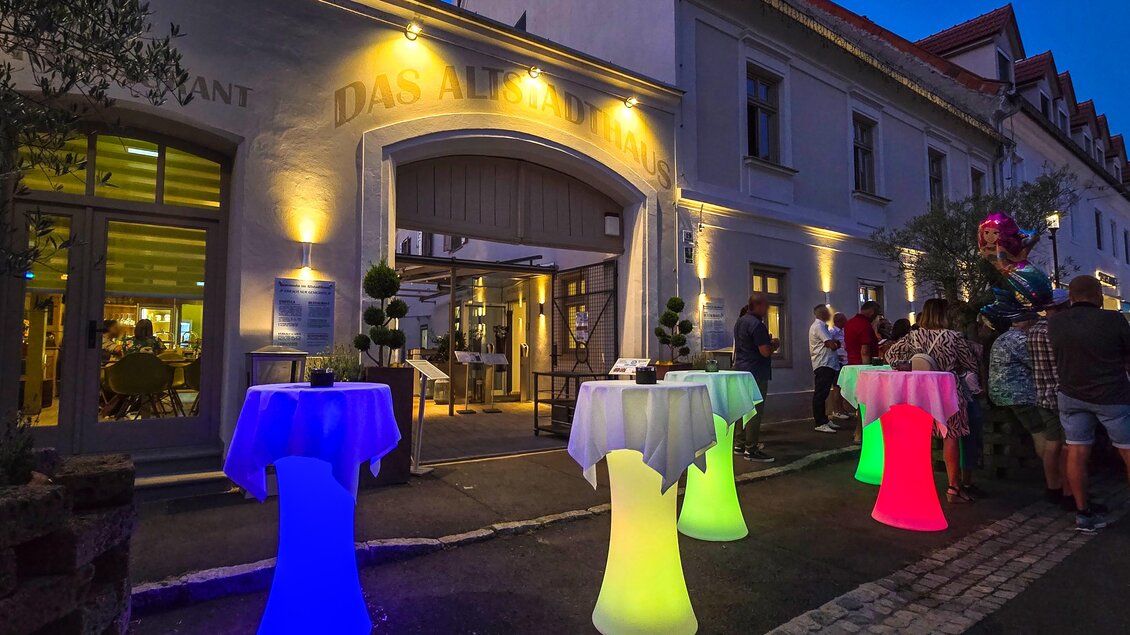 Ein Restaurant mit stilvollen, beleuchteten Stehtischen in verschiedenen Farben. Es ist Abend und Gäste stehen vor dem Eingang. | © © Das Altstadthaus