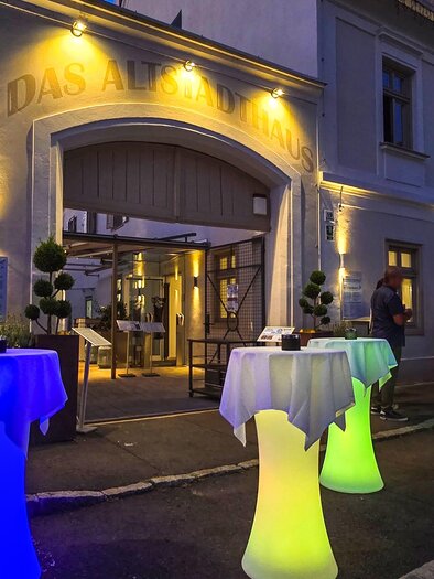 A restaurant with stylish, illuminated high tables in various colors. It is evening and guests are standing in front of the entrance. | © © Das Altstadthaus