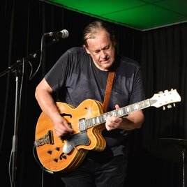 Ein Musiker spielt leidenschaftlich Gitarre auf der Bühne. Im Hintergrund sind Mikrofon und Schlagzeug sichtbar. | © @Christian Masser