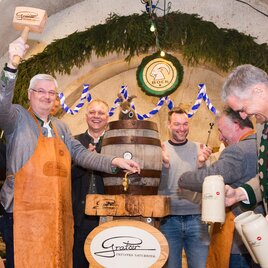 Eine Feier in einem Festsaal, bei der ein Bierfass angezapft wird. Die Teilnehmer tragen traditionelle Kleidung und zeigen Freude. | © Matthias Pöltl