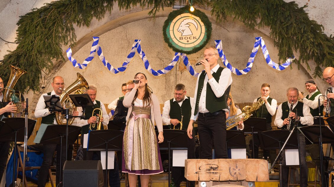 Eine Blasmusikgruppe tritt auf einer festlich geschmückten Bühne auf. Zwei Sänger stehen im Vordergrund und präsentieren ein musikalisches Stück.