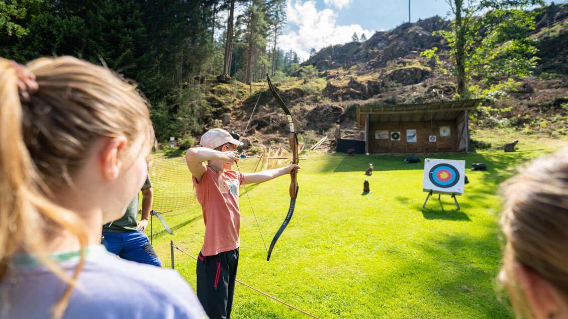 Ein Jugendlicher steht mit einem Bogen im Freien und zielt auf eine Zielscheibe. Im Hintergrund sind Bäume und ein kleines Gebäude zu sehen. | © TVB/Laura Passenbrunner