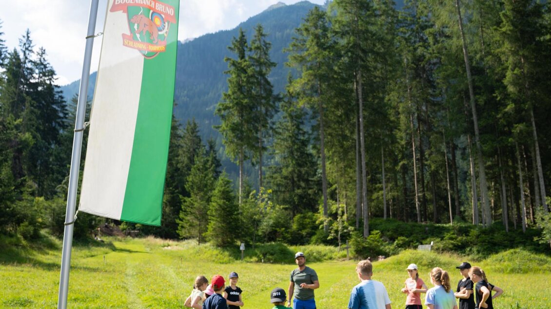 Eine Gruppe von Kindern steht auf einer grünen Wiese unter einem blauen Himmel. Im Hintergrund sind Wälder und Berge sichtbar, und eine große Fahne weht neben ihnen. | © TVB/Laura Passenbrunner