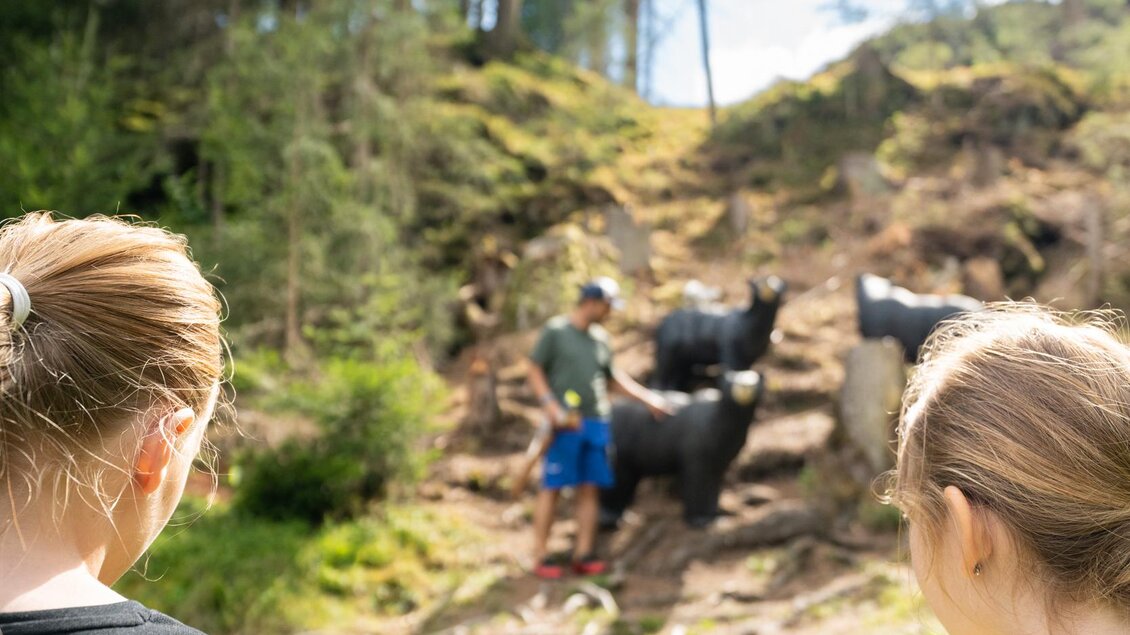 Zwei Kinder beobachten einen Mann, der mit Kühen in einer grünen Landschaft interagiert. Im Hintergrund sind Bäume und ein klarer Himmel zu sehen. | © TVB/Laura Passenbrunner