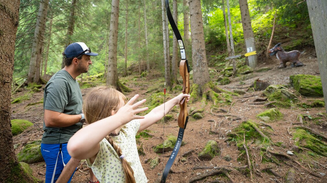 Ein Kind schießt mit einem Bogen auf ein Ziel in einem Wald. Ein Erwachsener beobachtet aufmerksam im Hintergrund. | © TVB/Laura Passenbrunner