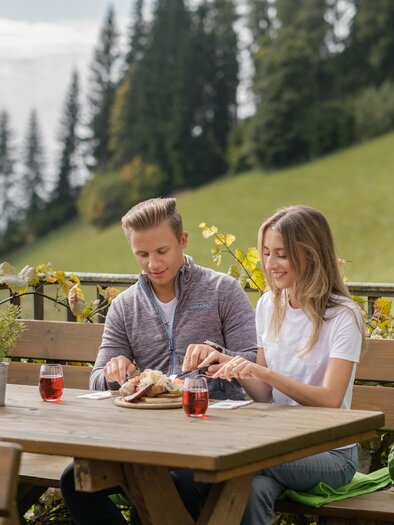 Ein Paar sitzt an einem Holztisch im Freien und genießt eine Mahlzeit. Im Hintergrund sind grüne Wiesen und Bäume zu sehen. | © STG / Elena Egger