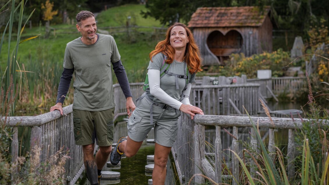 Ein Paar läuft auf einer Holzbrücke über einen Teich. Im Hintergrund sind grüne Wiesen und ein rustikales Häuschen zu sehen. | © STG/ Elena Egger