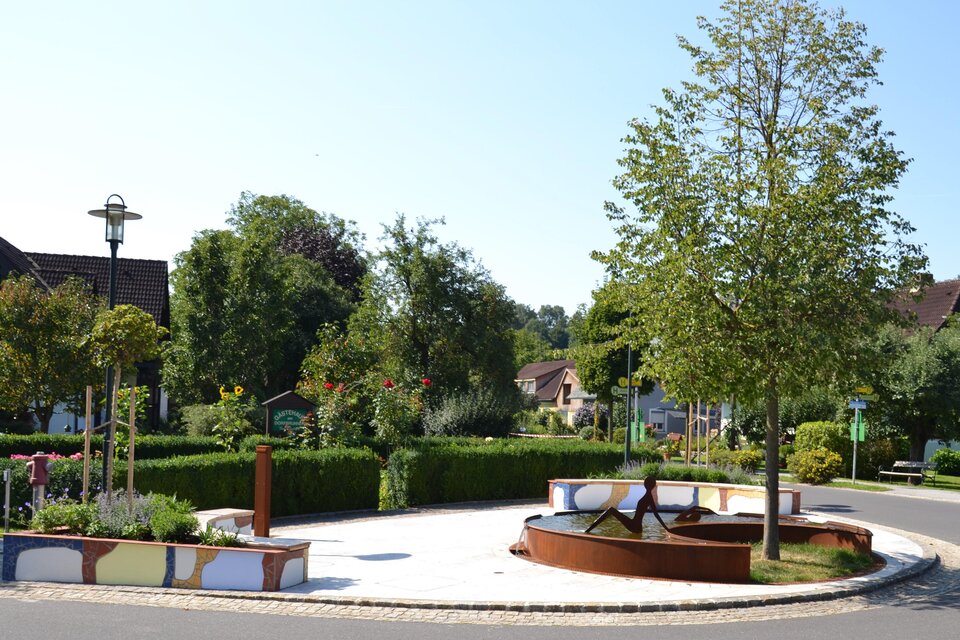 Ein sauberer Platz mit einem Brunnen und vielen grünen Bäumen. Umgeben von gepflegten Blumenbeeten und einer ruhigen Atmosphäre. | © © Kurkommission Bad Blumau