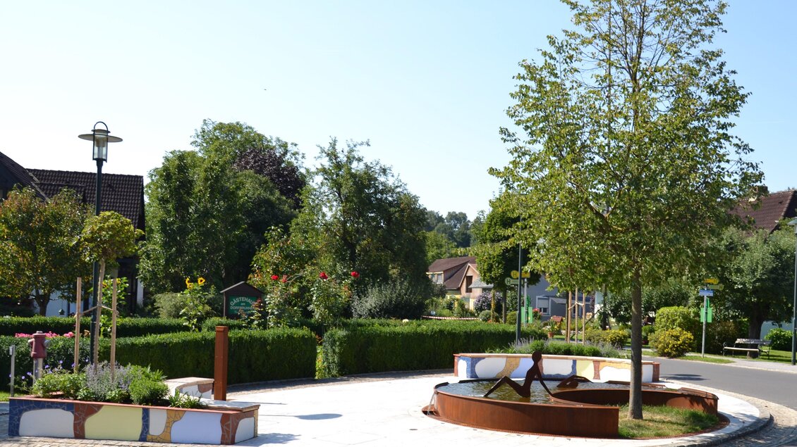 Ein sauberer Platz mit einem Brunnen und vielen grünen Bäumen. Umgeben von gepflegten Blumenbeeten und einer ruhigen Atmosphäre. | © © Kurkommission Bad Blumau