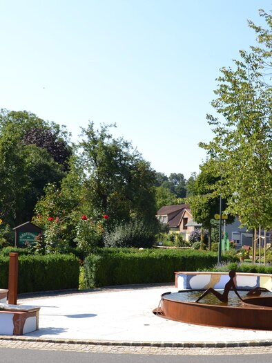 Ein sauberer Platz mit einem Brunnen und vielen grünen Bäumen. Umgeben von gepflegten Blumenbeeten und einer ruhigen Atmosphäre. | © © Kurkommission Bad Blumau