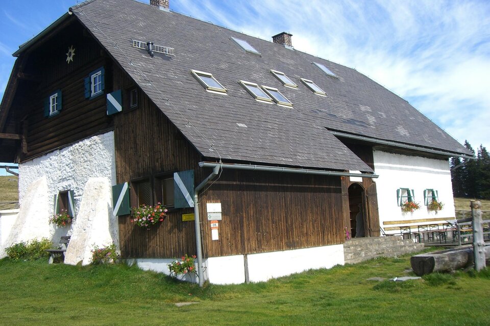 Ein traditionelles, zweigeschossiges Bauernhaus mit einem Holz- und Putz-Fassade. Das Gebäude befindet sich in einer ländlichen Umgebung mit grünem Gras und Fenster mit Blumen. | © Brendlhütte