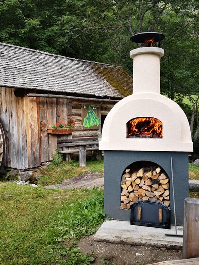 Ein schöner Außenofen aus Stein mit Holzlager steht in einem malerischen Garten. Im Hintergrund sieht man eine rustikale Holzscheune und grüne Bäume. | © Naturpark Sölktäler