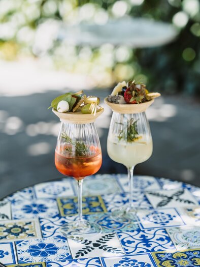 A cheerful woman in a colorful dress pours a drink into a glass. On the table, there are various foods and drinks, and in the background, you can see a green landscape. | © @Conny Leitgeb