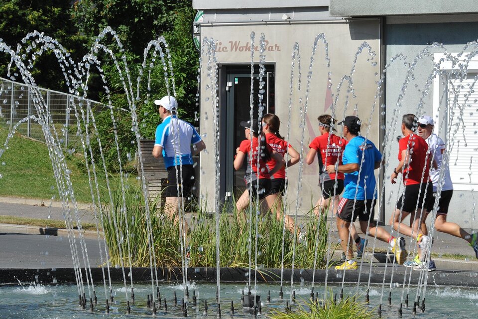 Eine Gruppe von Läufern joggt an einem Wasserspiel vorbei. Im Hintergrund sind moderne Gebäude und grünes Gras zu sehen. | © Thermen&Vulkanland