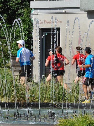 Eine Gruppe von Läufern joggt an einem Wasserspiel vorbei. Im Hintergrund sind moderne Gebäude und grünes Gras zu sehen. | © Thermen&Vulkanland