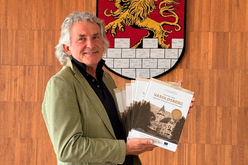 Bürgermeister Wolf-Maier präsentiert das Buch „Vasoldsberg“ vor dem Gemeindewappen. | © Kultur24