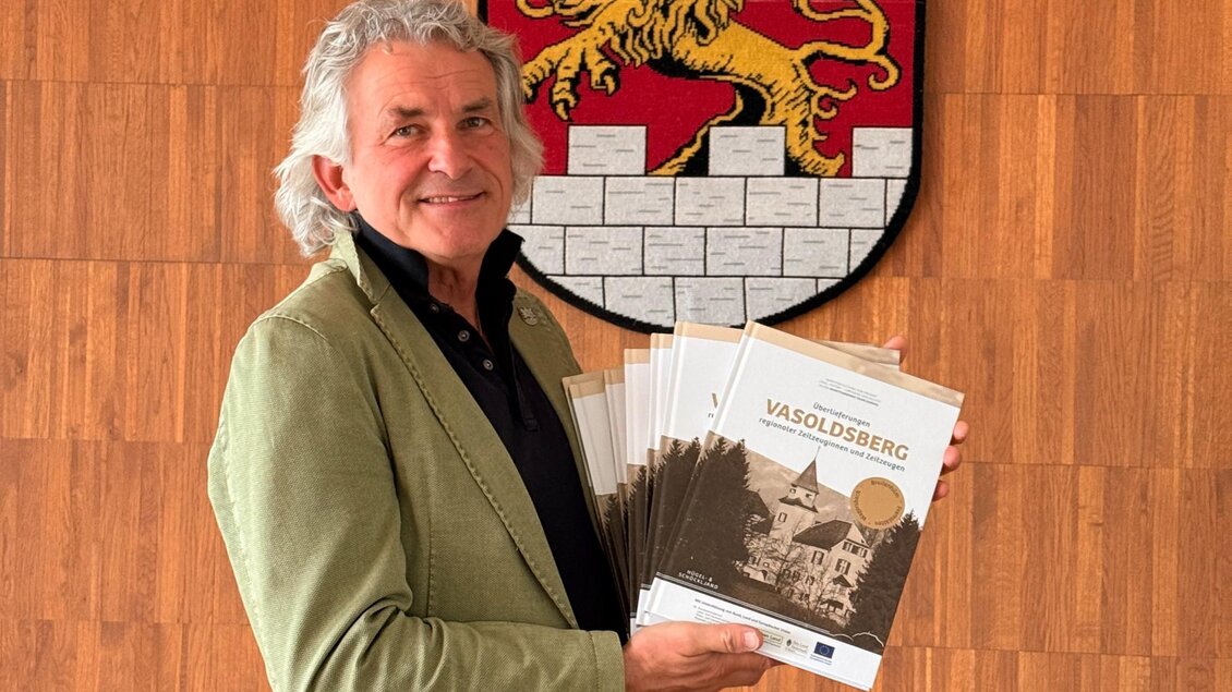 Bürgermeister Wolf-Maier präsentiert das Buch „Vasoldsberg“ vor dem Gemeindewappen. | © Kultur24