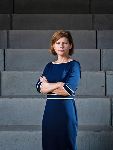 Eine Frau in einem blauen Kleid steht mit verschränkten Armen vor einer grauen Wand. Ihr Blick ist ernst und sie strahlt Selbstbewusstsein aus. | © Gianmaria Gava