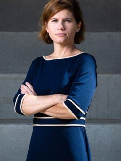 Eine Frau in einem blauen Kleid steht mit verschränkten Armen vor einer grauen Wand. Ihr Blick ist ernst und sie strahlt Selbstbewusstsein aus. | © Gianmaria Gava