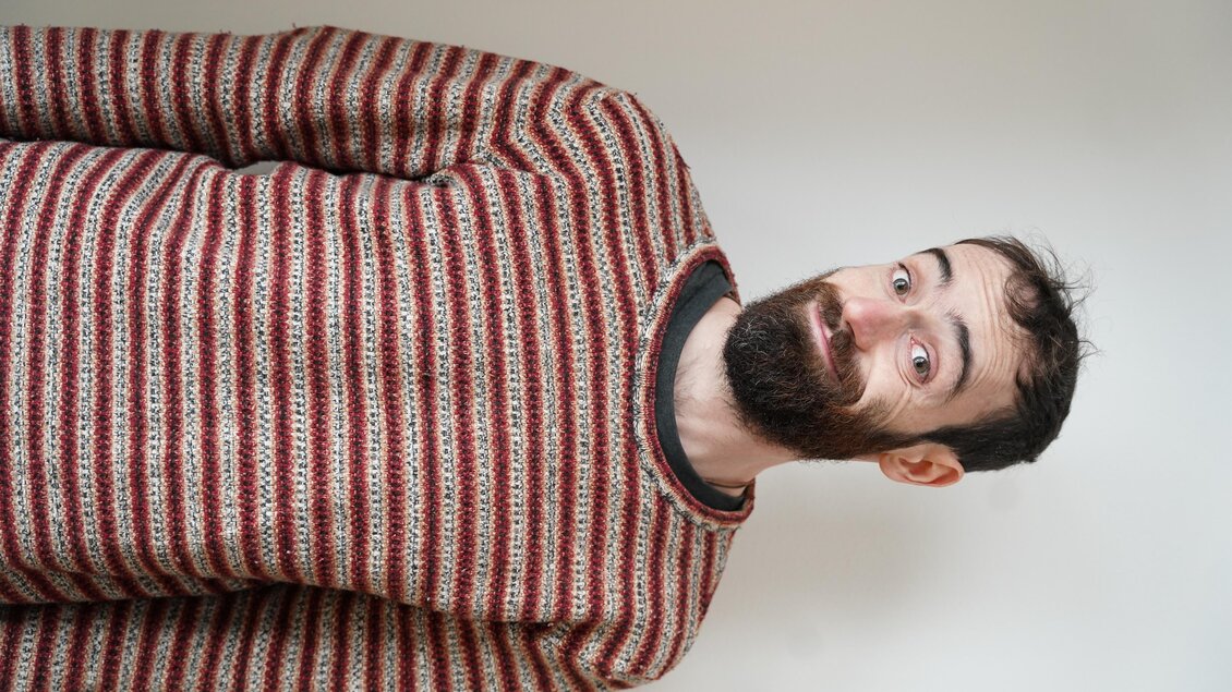 Ein Mann mit einem gestreiften Pullover steht vor einem neutralen Hintergrund. Er hat einen Bart und lächelt in die Kamera. | © Eva Plattner
