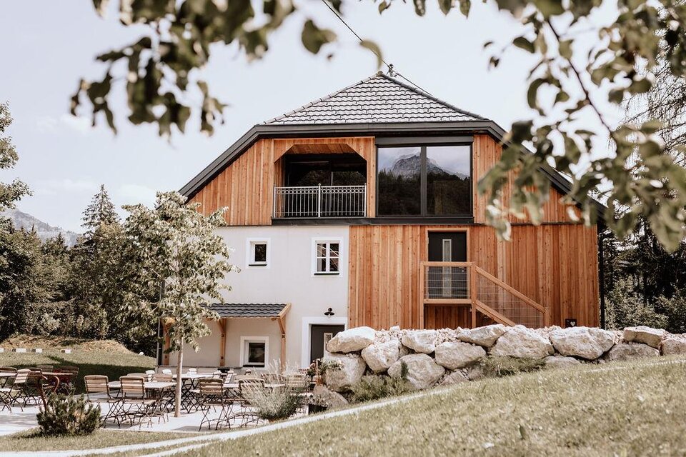 Das moderne Schutzhaus am hohen Camper mit Holzfassade und großen Fenstern befindet sich mittig vom Bild. Im Vordergrund sind Gartenmöbel und Grünflächen zu sehen.