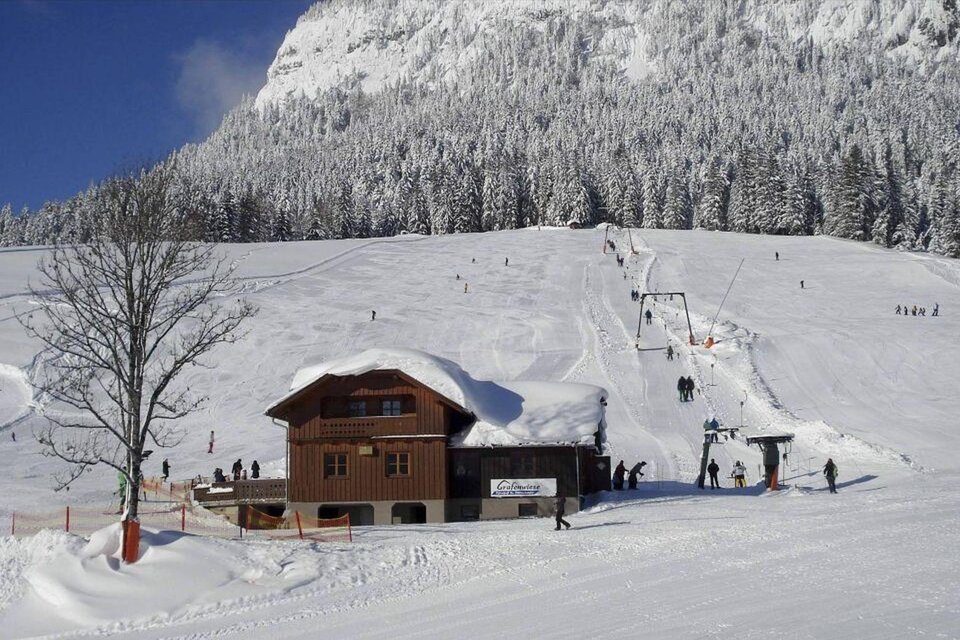 Ein schneebedecktes Skigebiet mit einem rustikalen Holzhaus. Im Hintergrund sind Wintersportler und eine Skipiste zu sehen. | © Grafenwiese