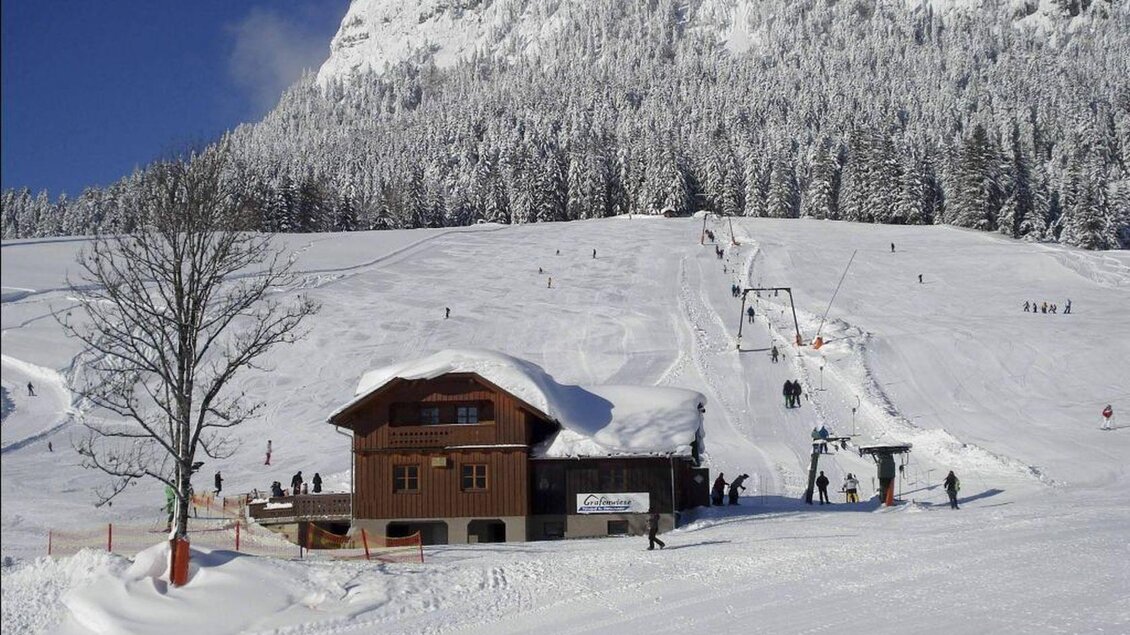 Ein schneebedecktes Skigebiet mit einem rustikalen Holzhaus. Im Hintergrund sind Wintersportler und eine Skipiste zu sehen. | © Grafenwiese
