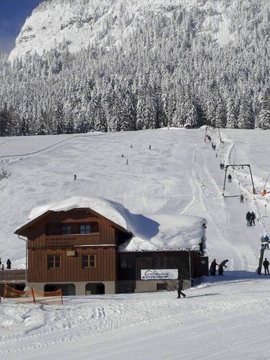 A snow-covered ski resort with a rustic wooden house. In the background, winter sports enthusiasts and a ski slope can be seen. | © Grafenwiese