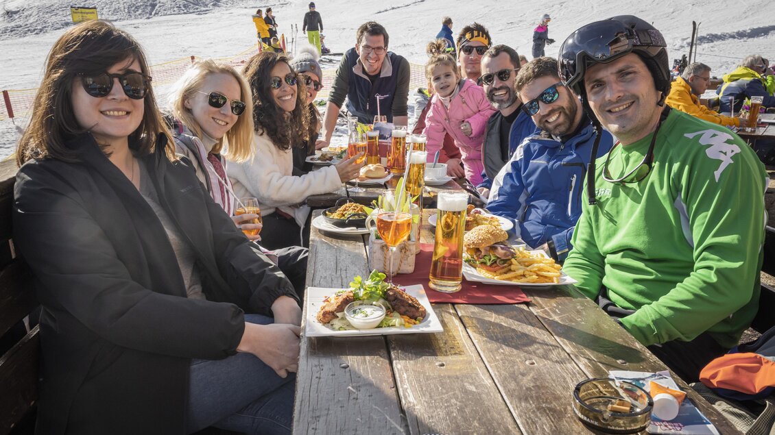 Eine Gruppe von Menschen sitzt an einem Tisch in einem Skilager und genießt Essen und Getränke. Im Hintergrund sind schneebedeckte Berge zu sehen. | © Melanie Fritz