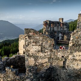 Eine historische Burgruine auf einem Hügel, umgeben von grünen Wäldern und Bergen. Im Vordergrund sitzen zwei Personen auf einer Mauer und genießen die Aussicht. | © TV Murau