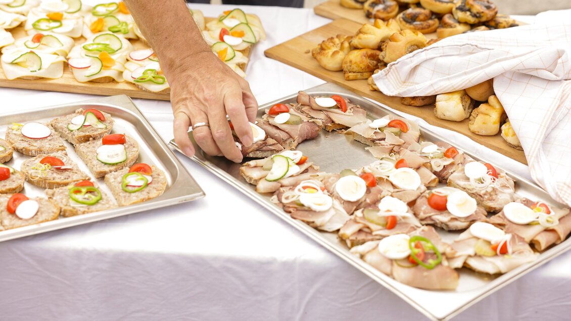 Eine Person legt Lebensmittel auf eine silberne Platte mit verschiedenen belegten Broten. Im Hintergrund sind unterschiedliche Snacks und Brötchen zu sehen. | © Gemeinde Bad Loipersdorf