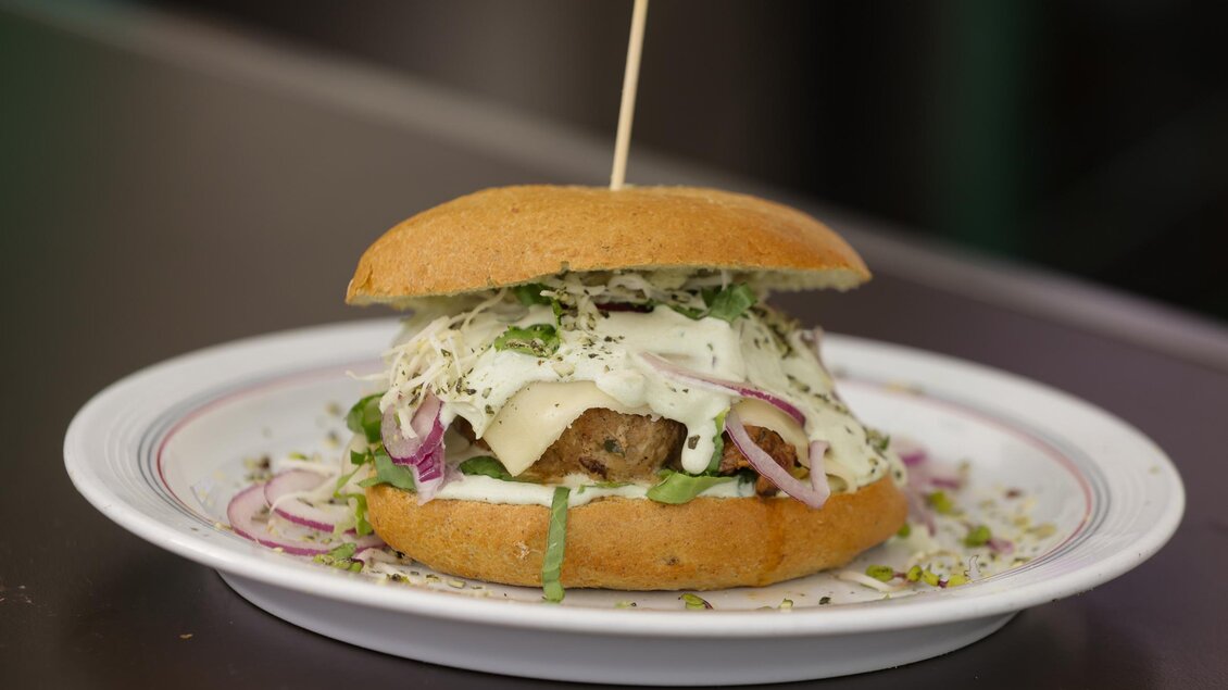 Ein leckerer Burger mit einer goldenen Brötchenoberseite, gefüllt mit Fleisch, frischem Gemüse und einer cremigen Sauce. Er ist ansprechend angerichtet auf einem weißen Teller. | © Gemeinde Bad Loipersdorf