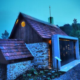 Ein modernes Haus mit Holz- und Steinelementen, das von einem sanften blauen Licht erleuchtet wird. Die Architektur kombiniert traditionelle und zeitgenössische Stile. | © Die Amtmann