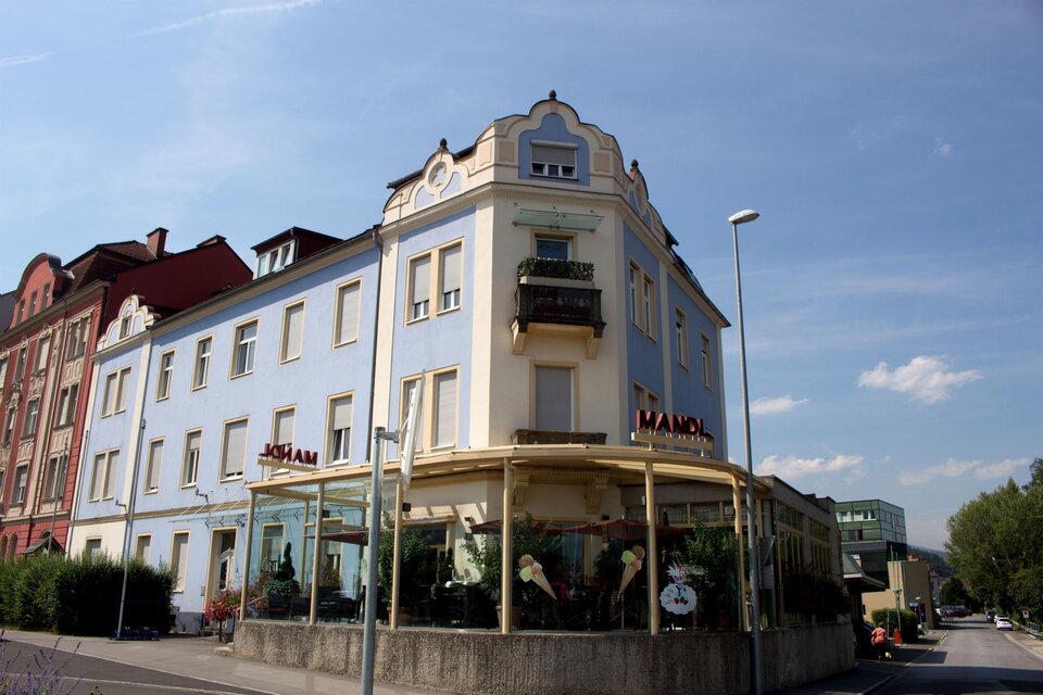 Ein elegantes, blaues Gebäude mit einem schönen Erker und einer großen Glasfassade. Das Hotel hat einen einladenden Eingang und liegt an einer Straßenkreuzung. | © C.Stein