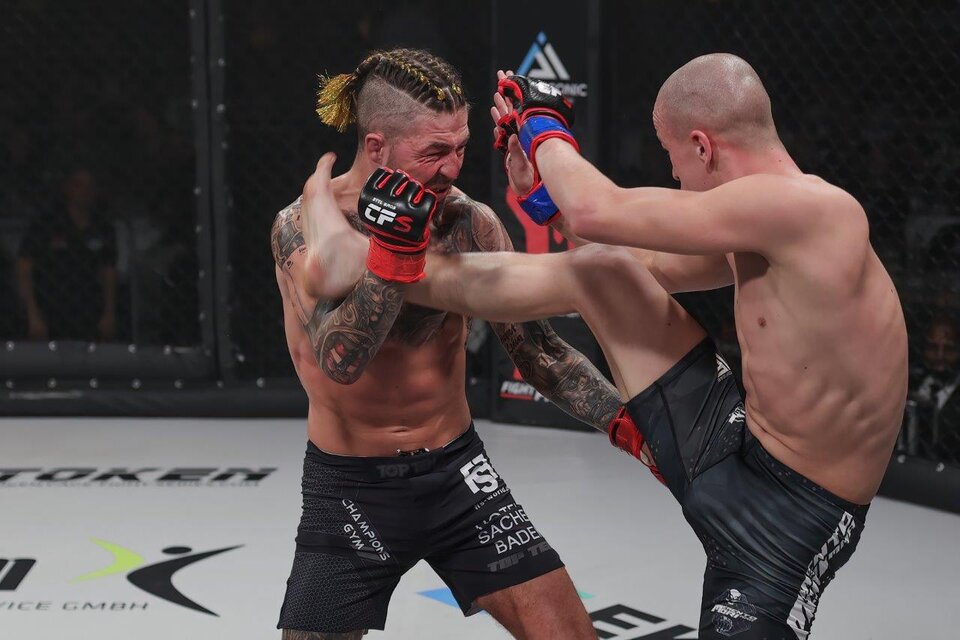 Zwei Kämpfer im Oktagon während eines MMA-Matches. Der Kämpfer mit geflochtenem Haar trifft seinen Gegner mit einem hohen Kick. | © Bobipix