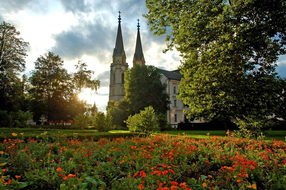 Ein schöner Garten mit bunten Blumen und grünen Bäumen. Im Hintergrund sind hohe Kirchtürme und ein bewölkter Himmel zu sehen. | © Thomas Sattler
